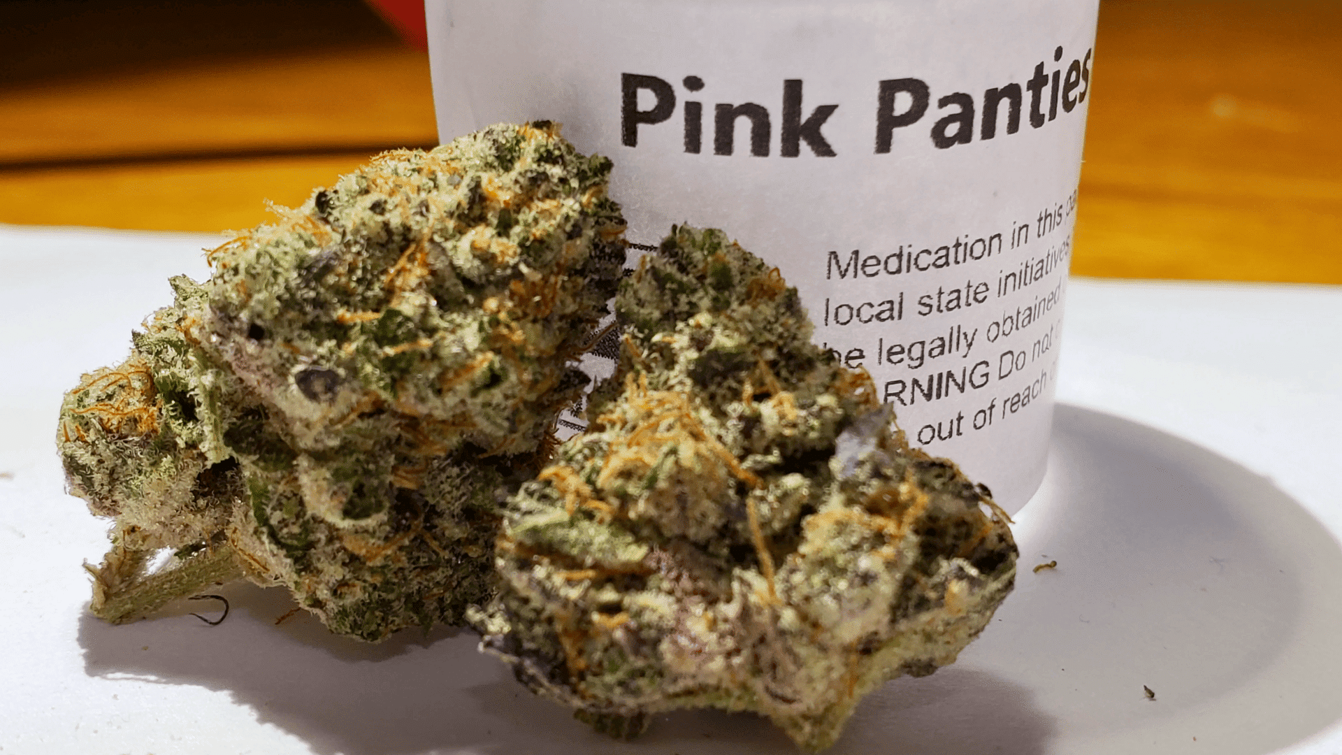 pink panties cannabis buds with light green leaves and orange hairs next to a white medical container on a white surface