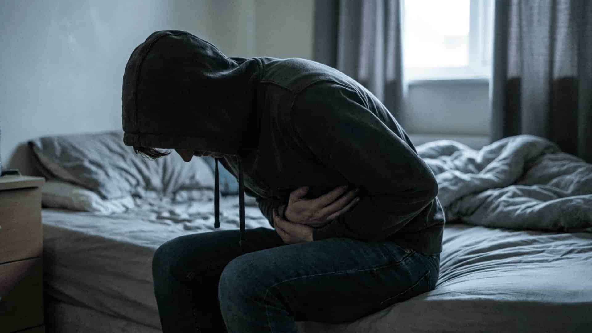 person hunched over on a bed in a dimly lit room with hands pressed against their stomach in pain