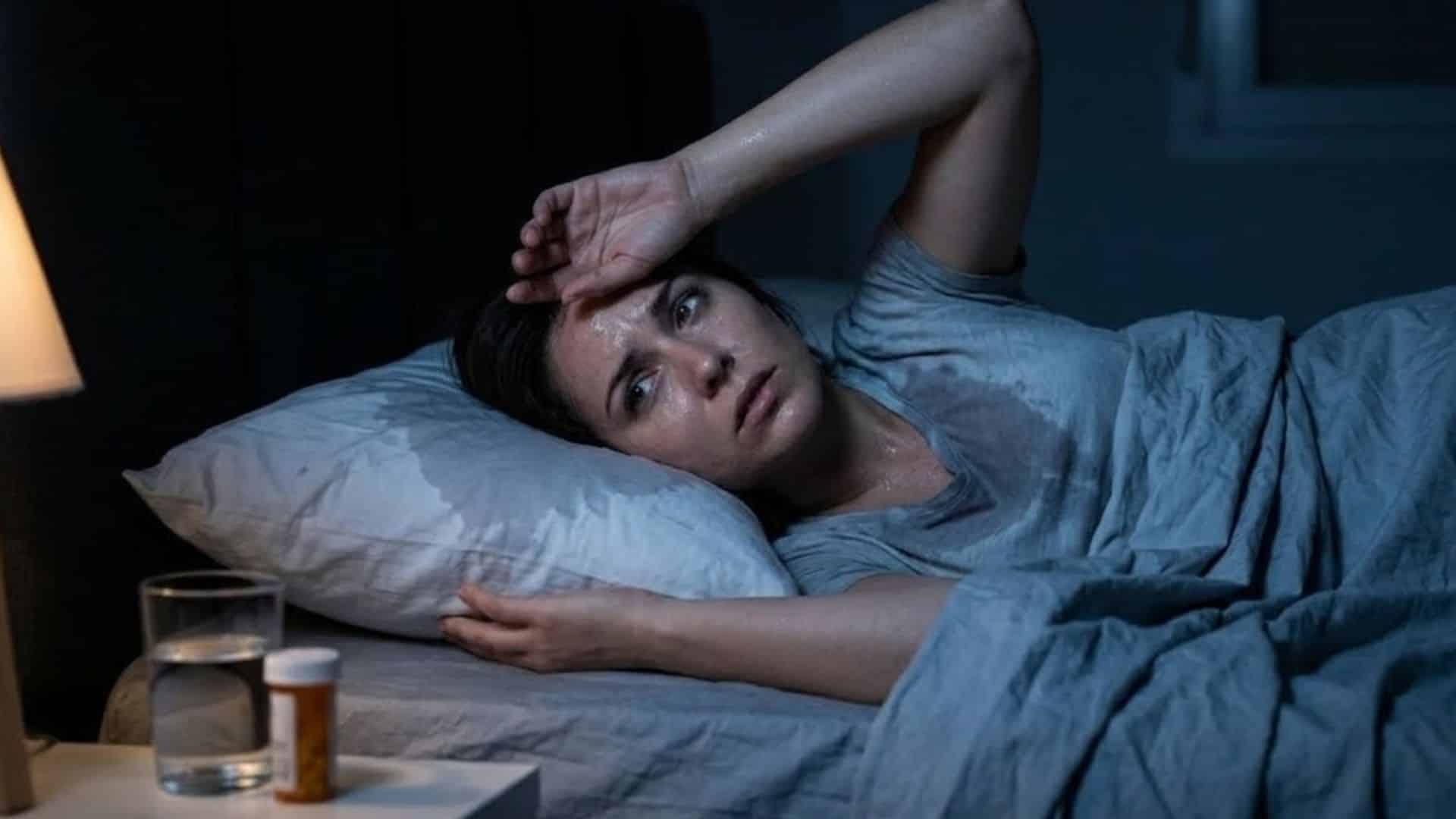 woman lies awake in a dark bedroom, visibly sweating with a damp pillow and shirt, looking distressed and uncomfortable