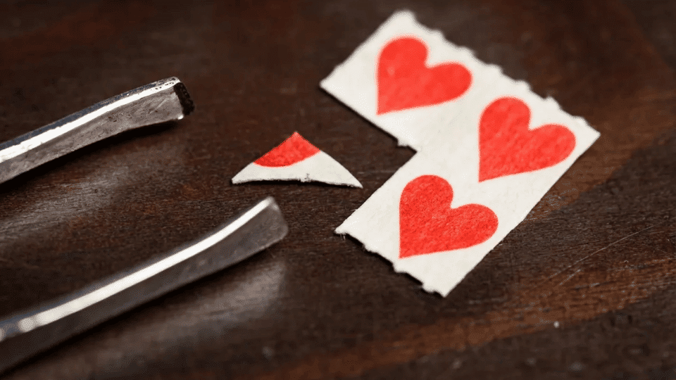 torn playing cards with red hearts on dark wooden surface with tweezers close up