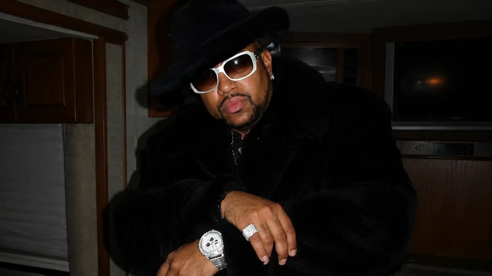 a man in a black fur coat and hat, wearing oversized sunglasses and jewelry, striking a confident pose in a dim room