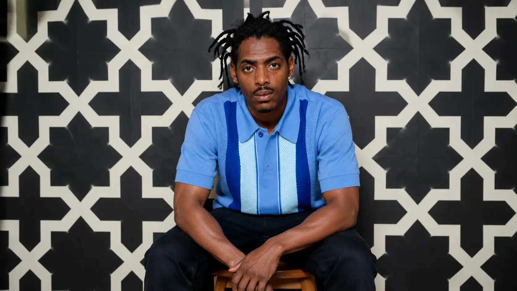 a man in a blue polo shirt sits on a wooden stool in front of a black-and-white geometric patterned background