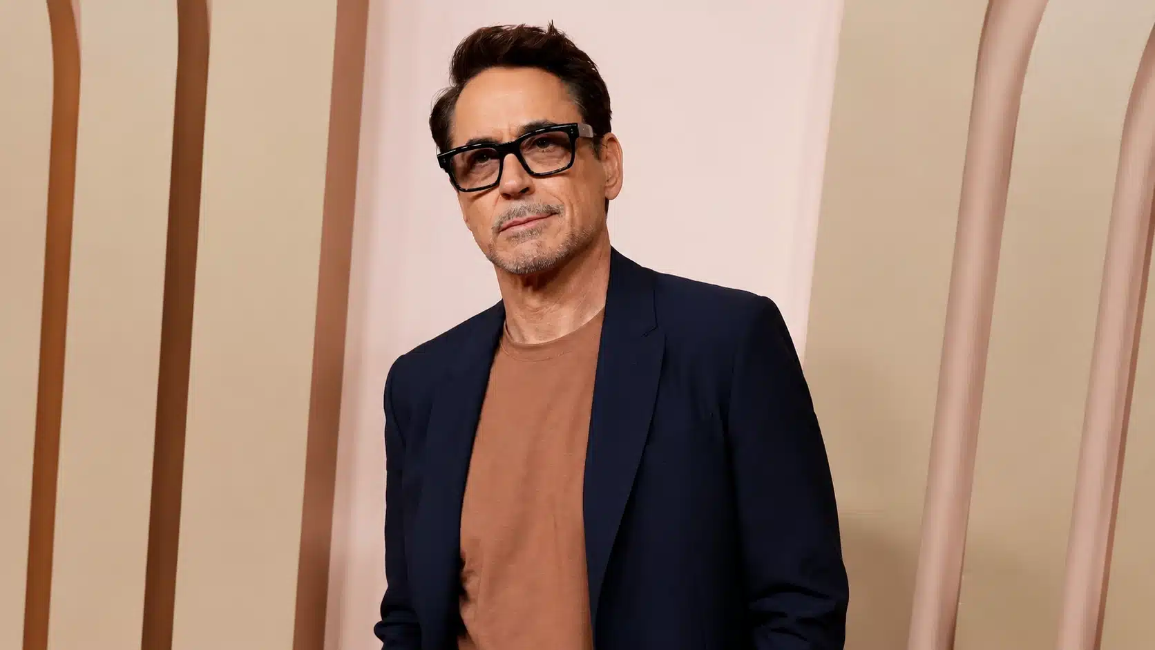 a man in a navy blazer and tan shirt, with black glasses, stands in front of a warm-toned backdrop