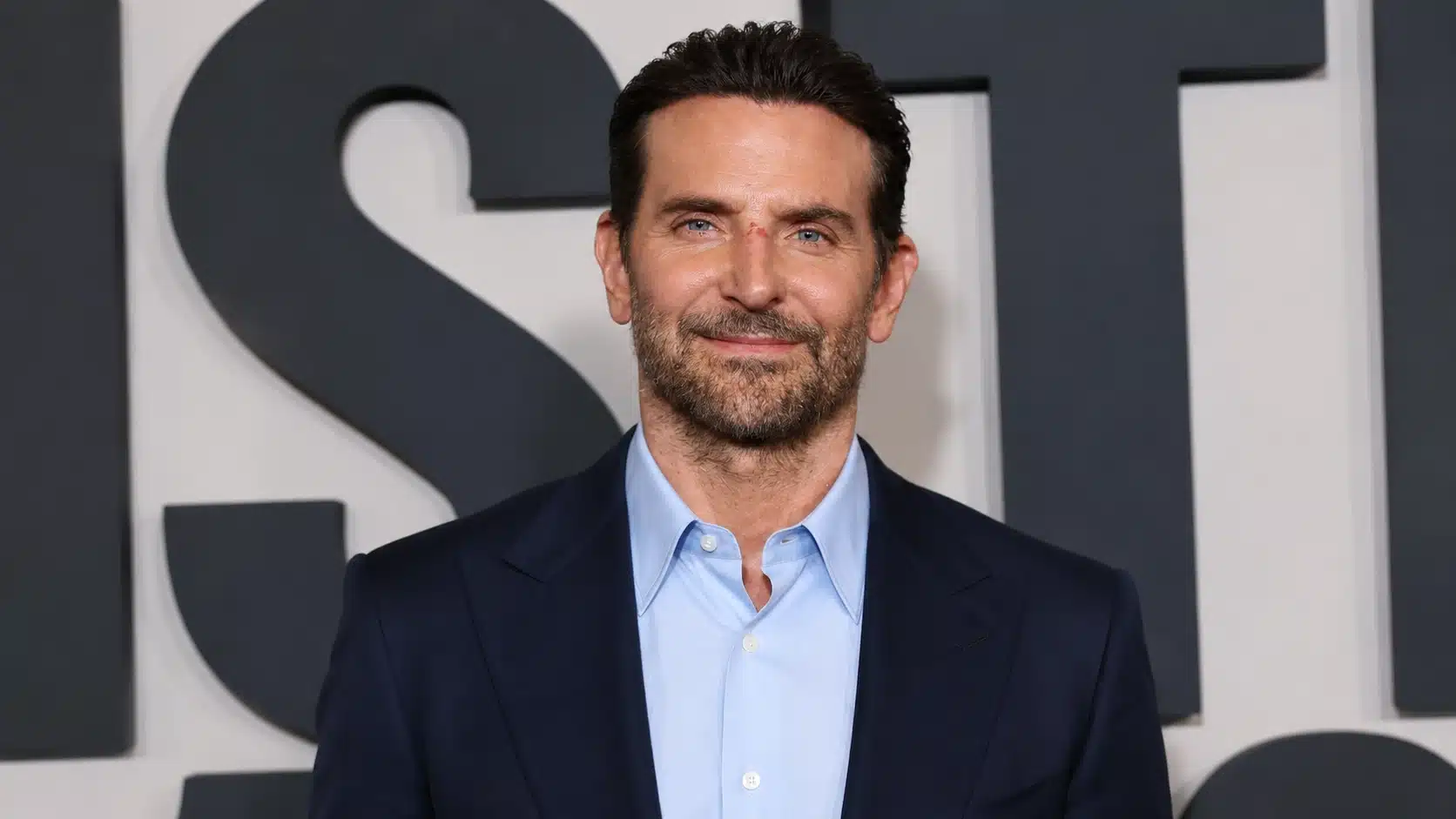 a man in a navy blue suit and light blue shirt smiles confidently against a dark logo-like background