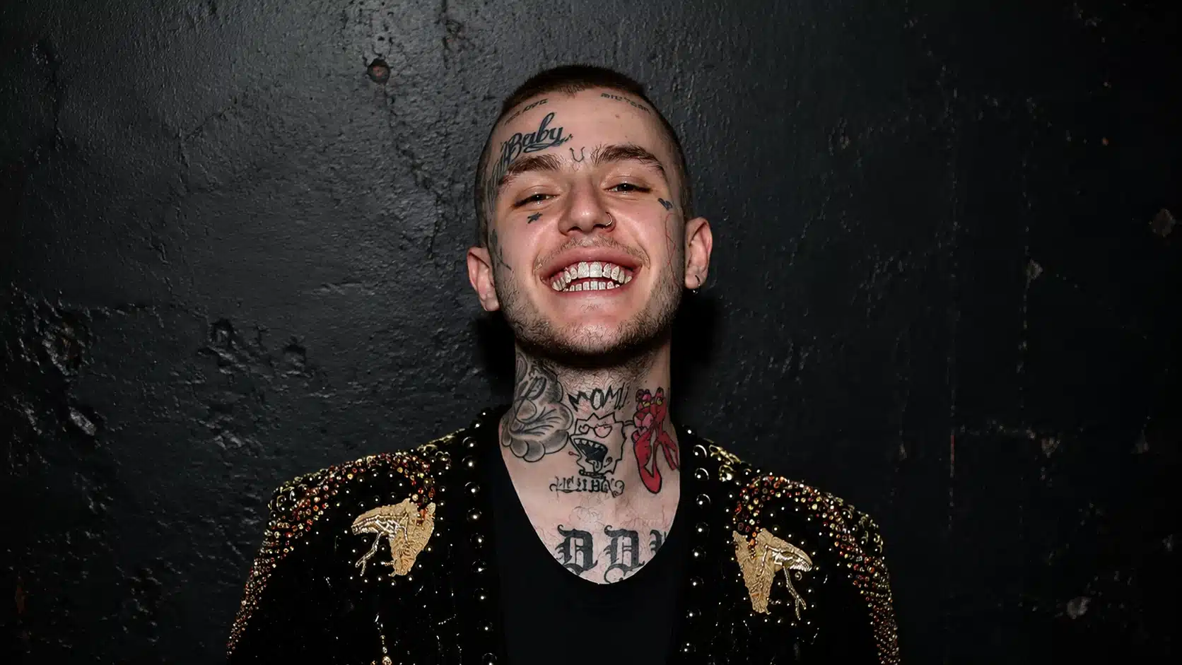 a man smiling confidently in a studded jacket with tattoos visible on his neck and face, against a black wall