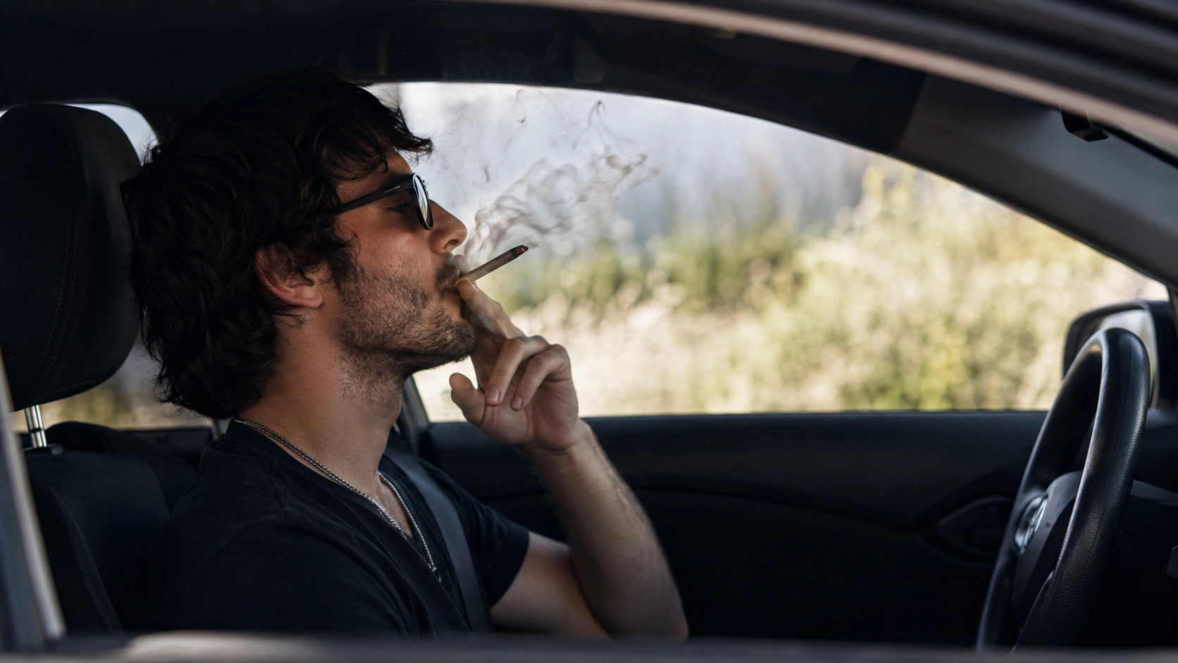 a side-angle view shows a man sitting in a car, smoking a cannabis joint as smoke drifts near his face