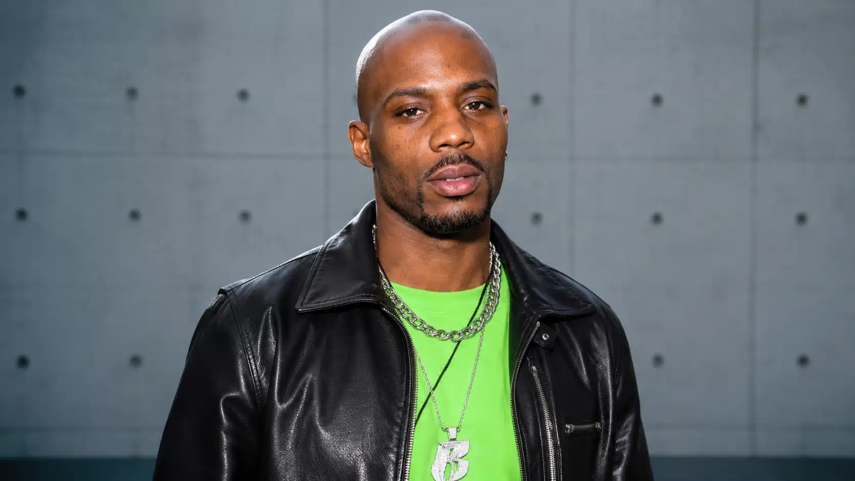 a man wearing a black leather jacket and neon green shirt with silver chains, standing against a concrete wall outdoors