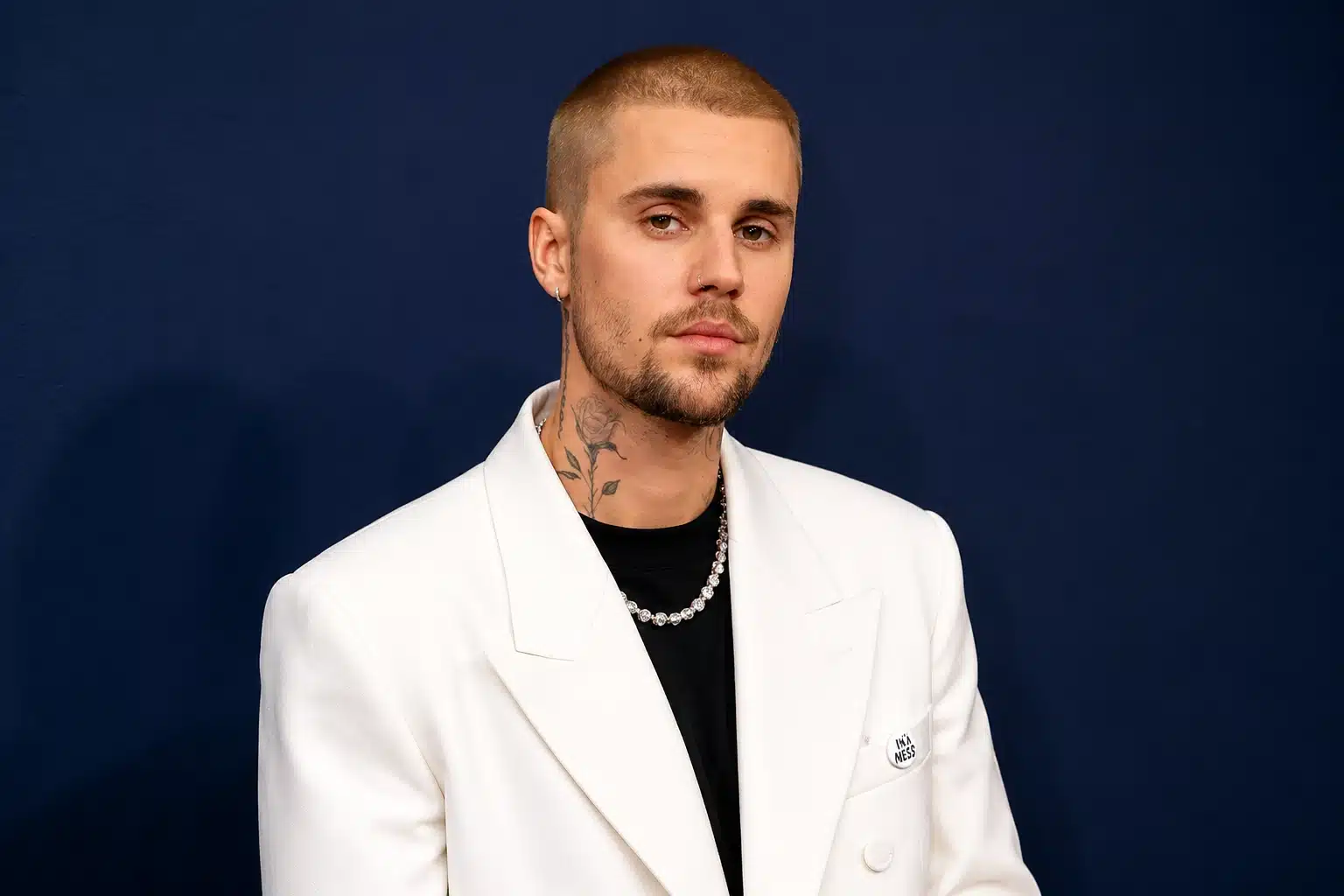 a man with a buzz cut and tattoos, wearing a white blazer and black shirt, stands against a dark navy background