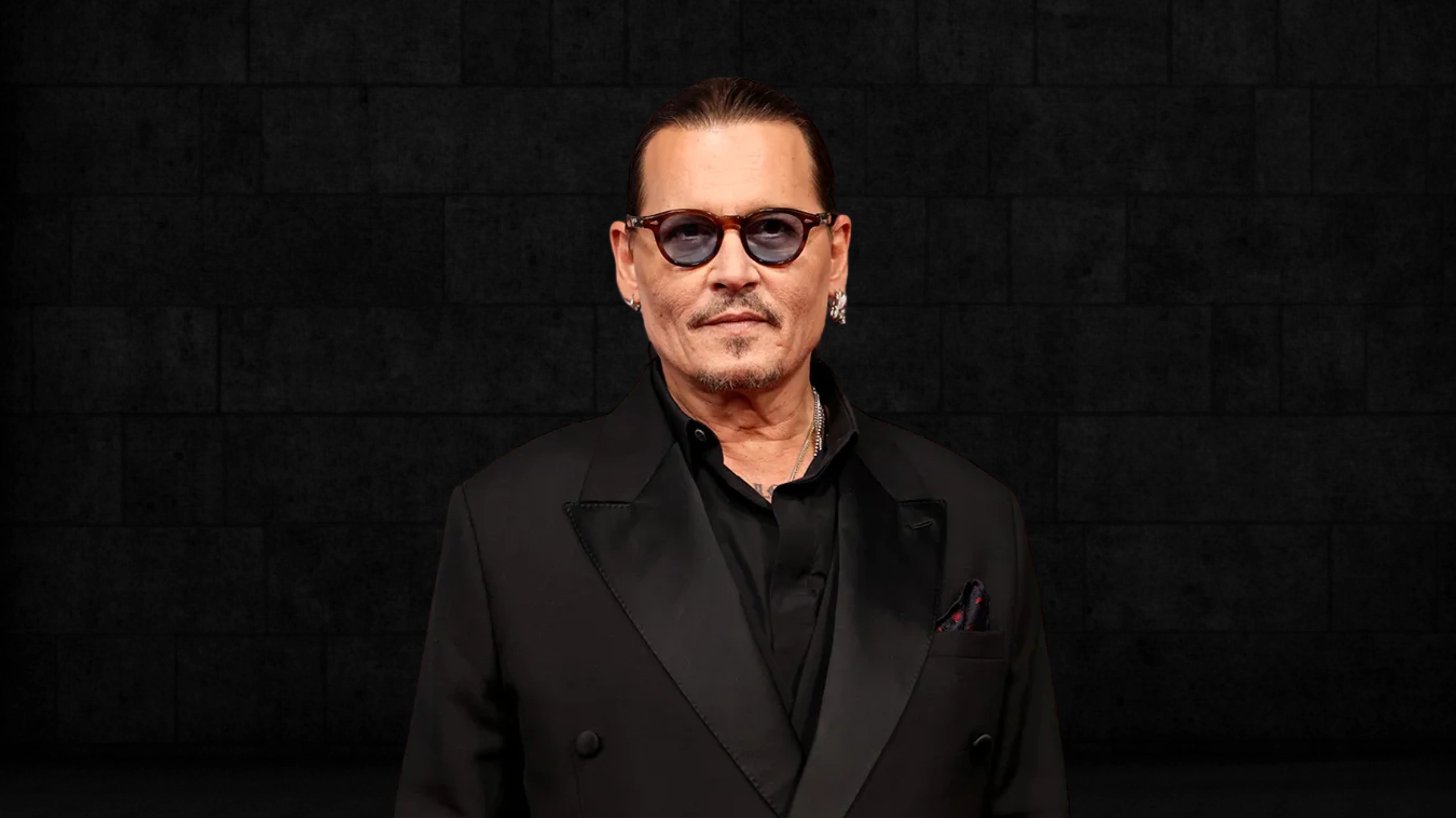 a man with a mustache and sunglasses, wearing a black suit and shirt, stands confidently in front of a dark stone wall