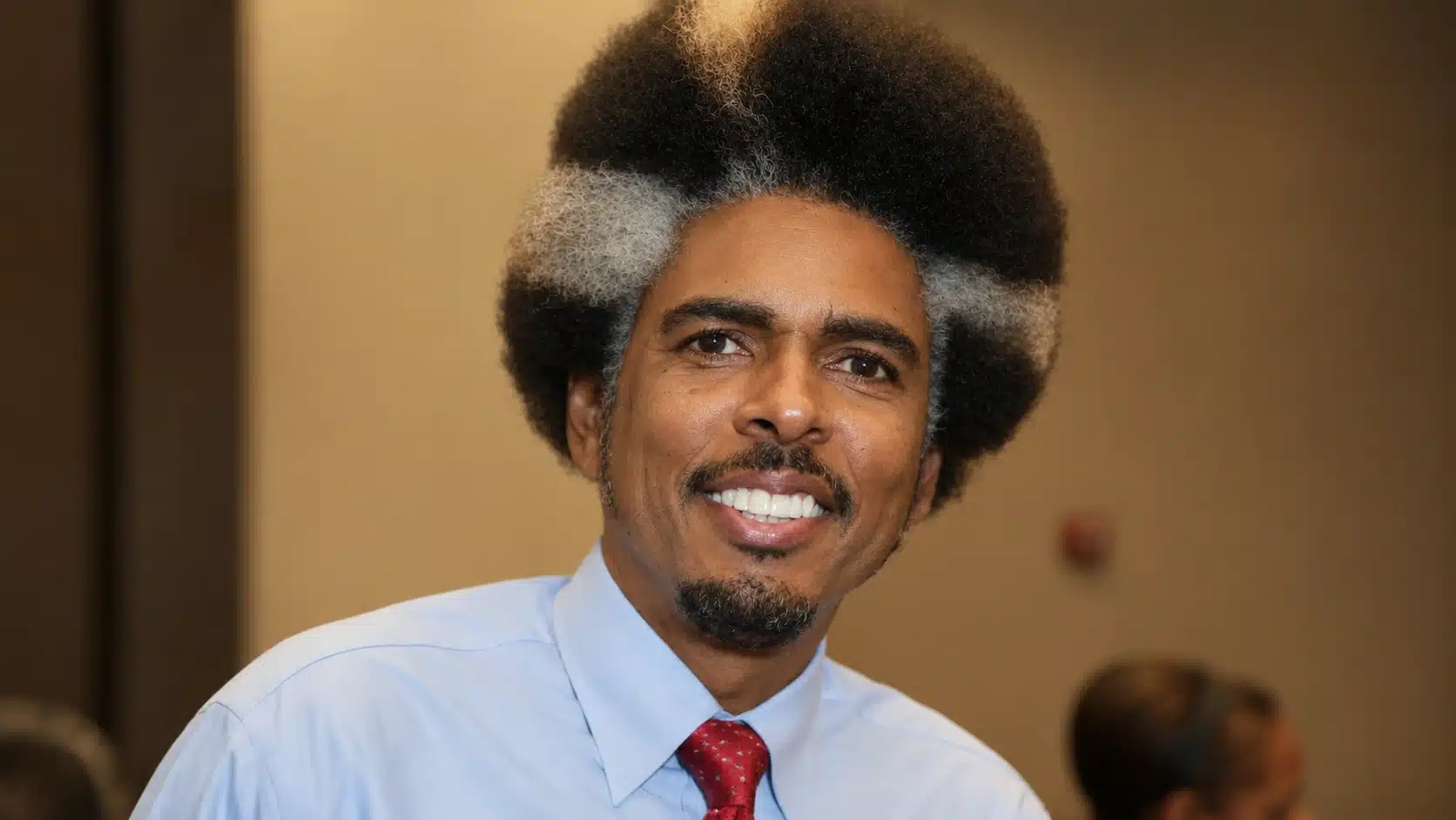 a man with a unique two-tone afro, wearing a light blue shirt and red tie, smiling warmly against a soft background