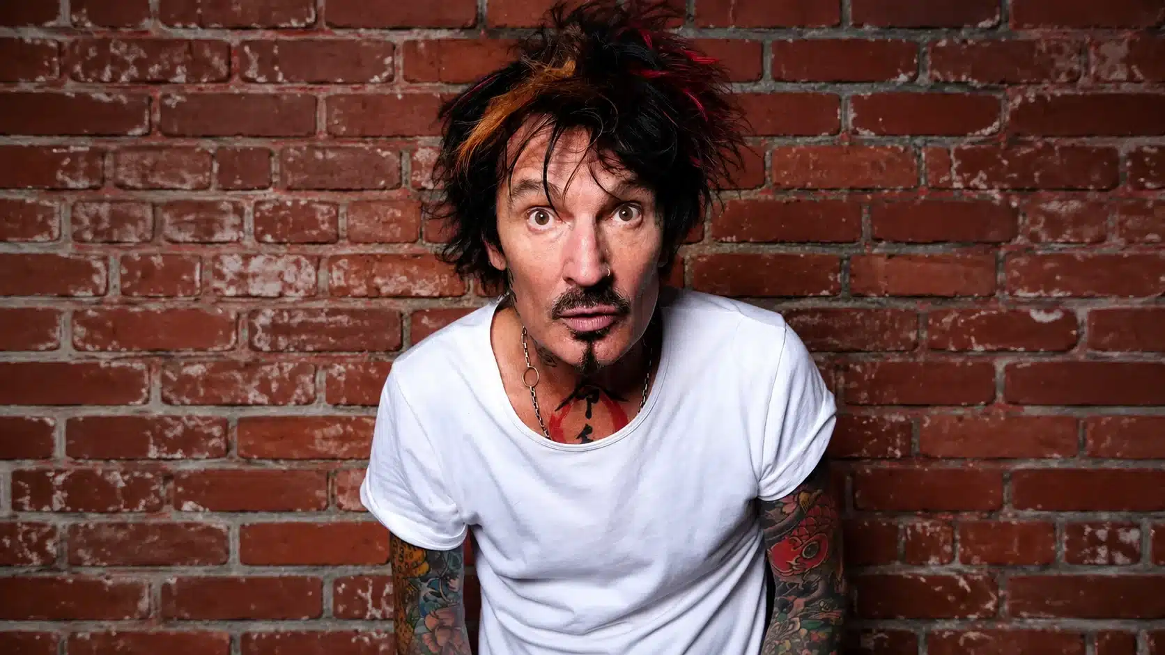 a man with colorful hair and tattoos, wearing a white t-shirt, stands in front of a red brick wall