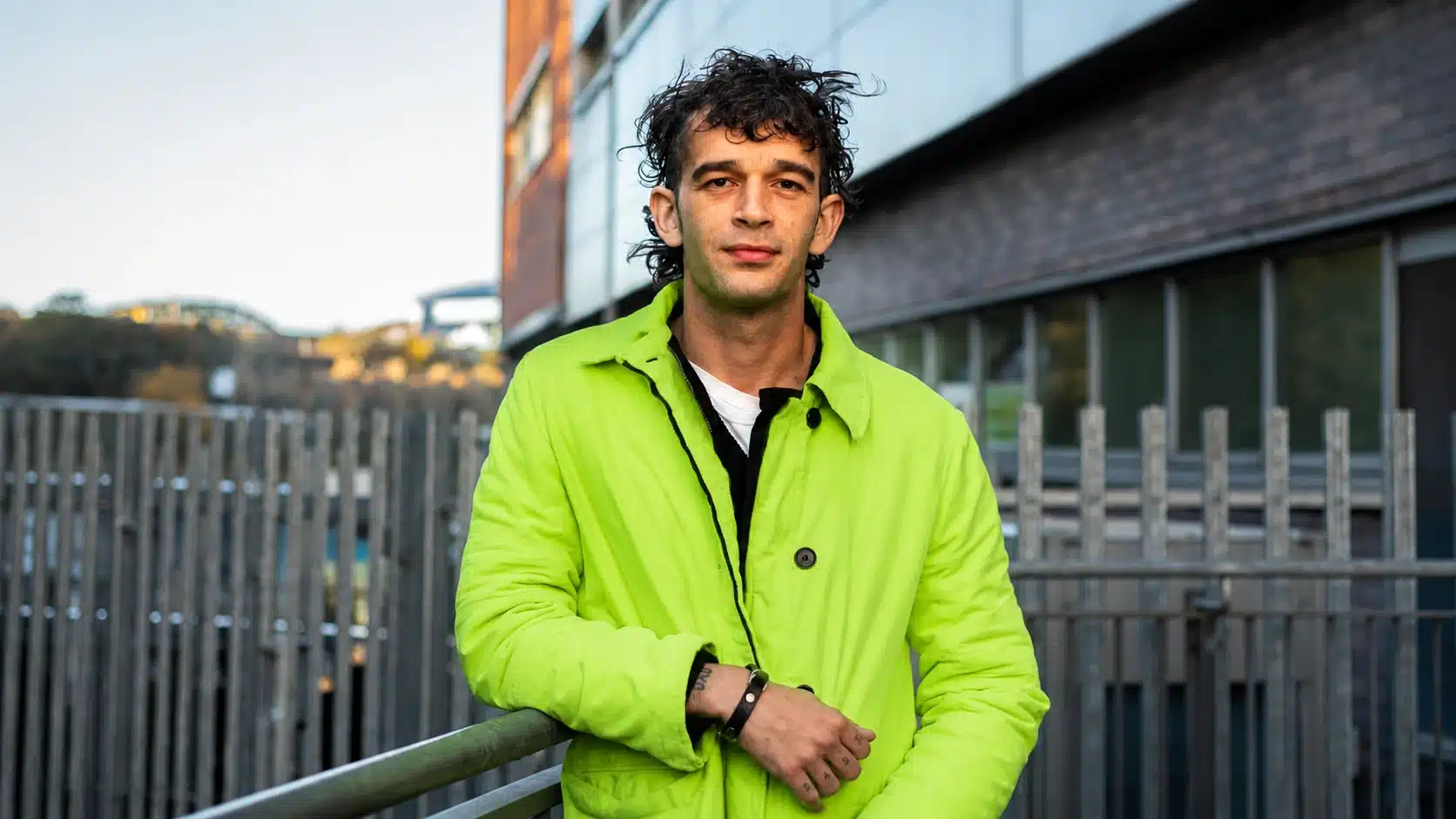 a man with curly hair, wearing a neon green jacket, poses casually outdoors, leaning on a metal railing