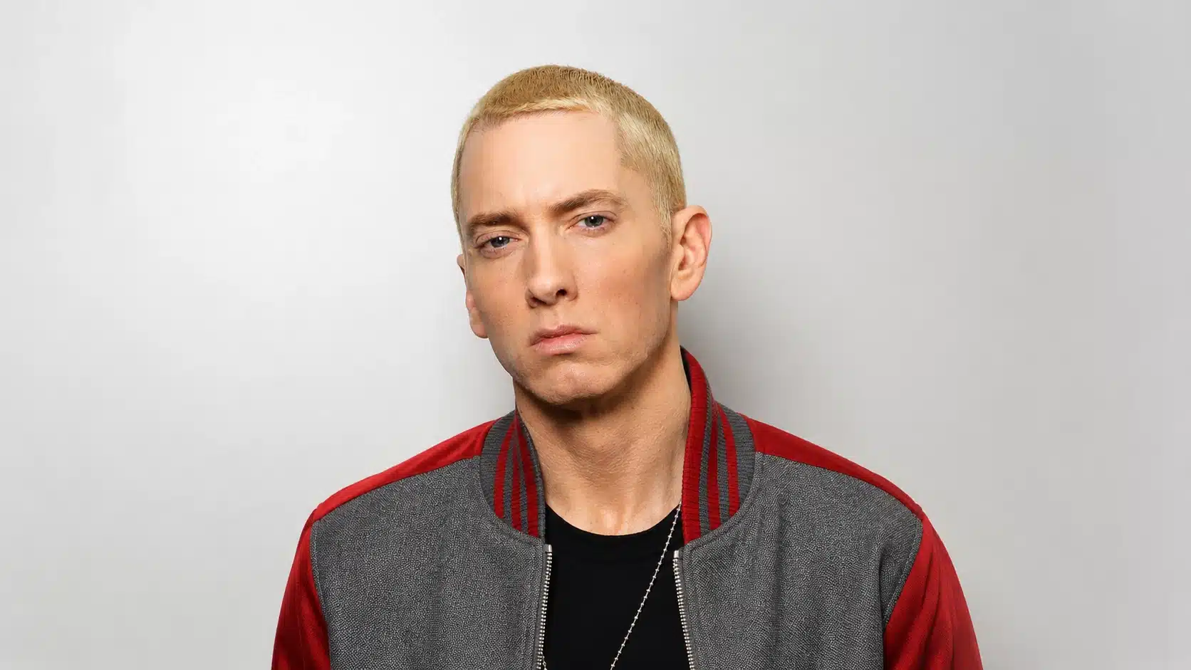 a man with light blond hair and a serious expression, wearing a gray and red bomber jacket against a neutral background