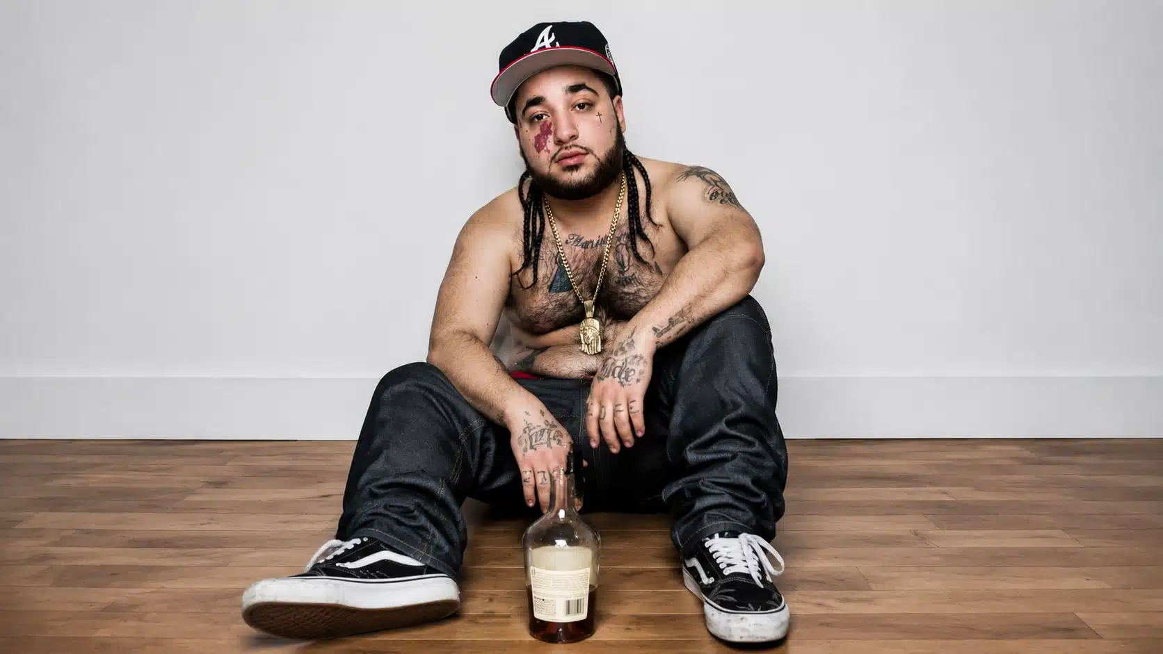 a man with tattoos and braids sitting on the floor in a casual pose, with a liquor bottle in front
