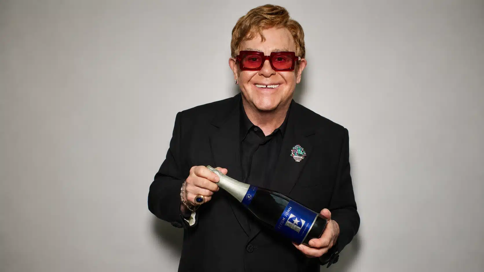 a smiling man in a black suit, wearing red glasses, holding a bottle of champagne against a gray background