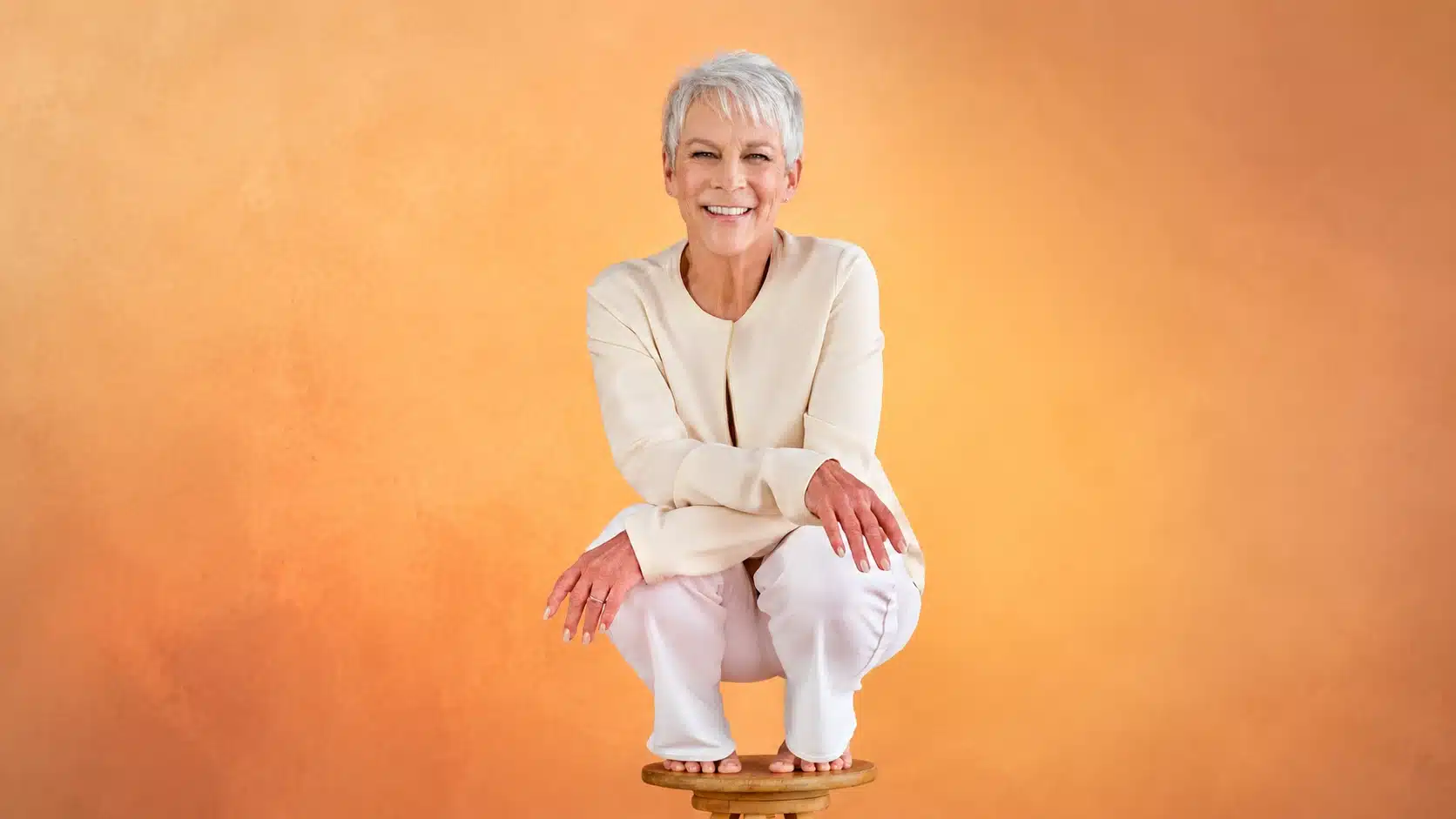 a smiling older woman with short gray hair squats on a wooden stool, dressed in cream and white, against a sunset wall