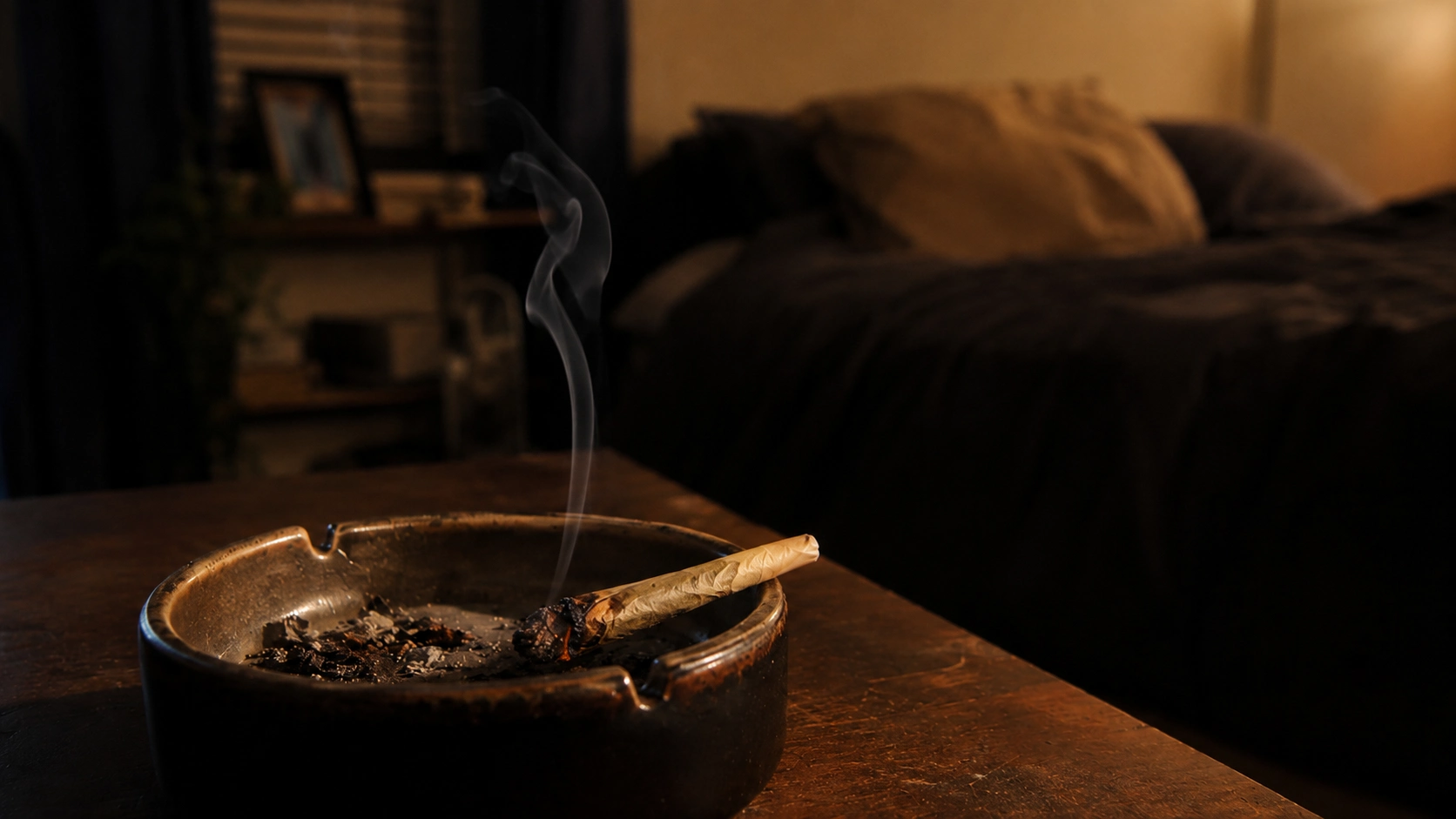 a lit cannabis joint smolders in a metal ashtray on a wooden table, with smoke rising in a dim bedroom
