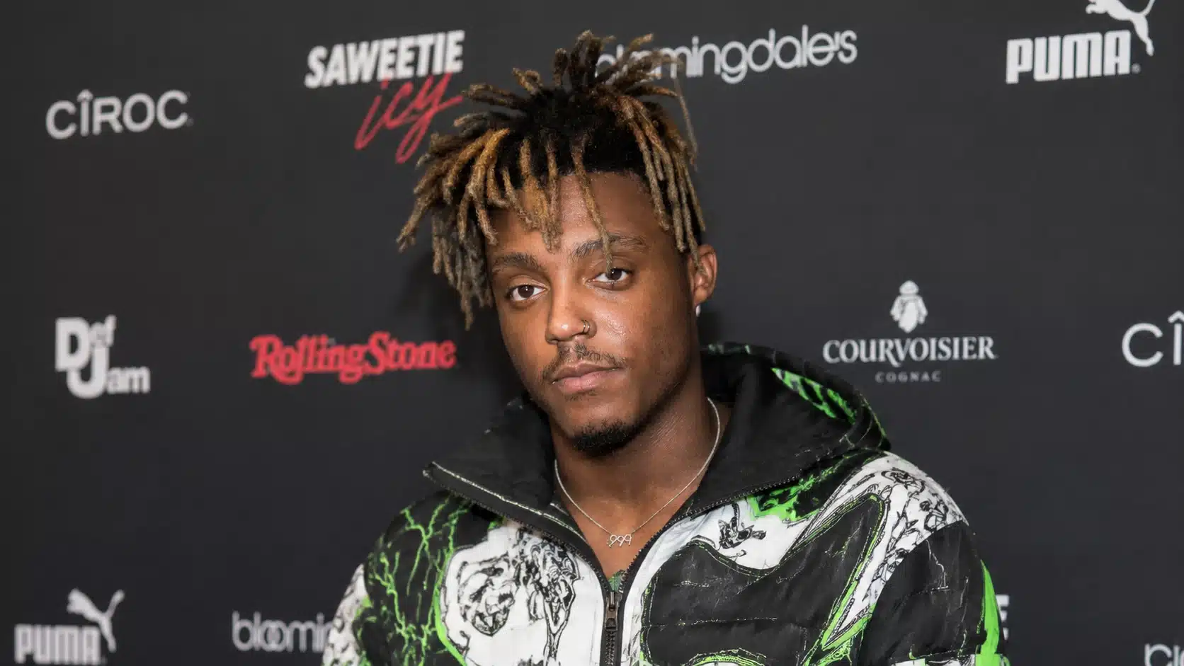 a tattooed man wearing a black, white, and neon green jacket with a 999 necklace, in front of a brand logo backdrop