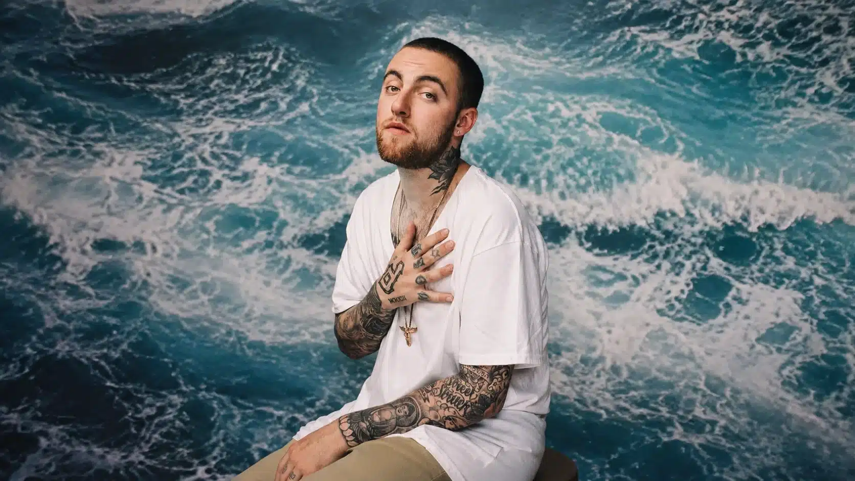 a tattooed man wearing a white t-shirt sits on a stool in front of an ocean-patterned backdrop, striking a calm pose