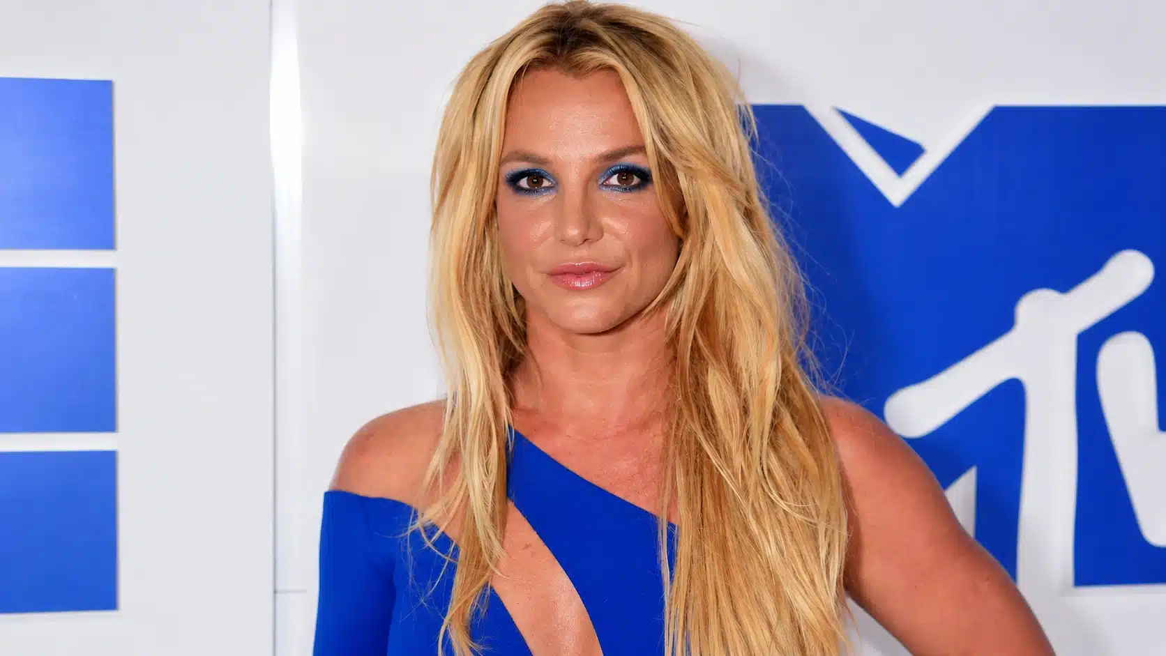 a woman with blonde hair, wearing blue eyeshadow and a cobalt blue dress, poses confidently in front of a mtv backdrop