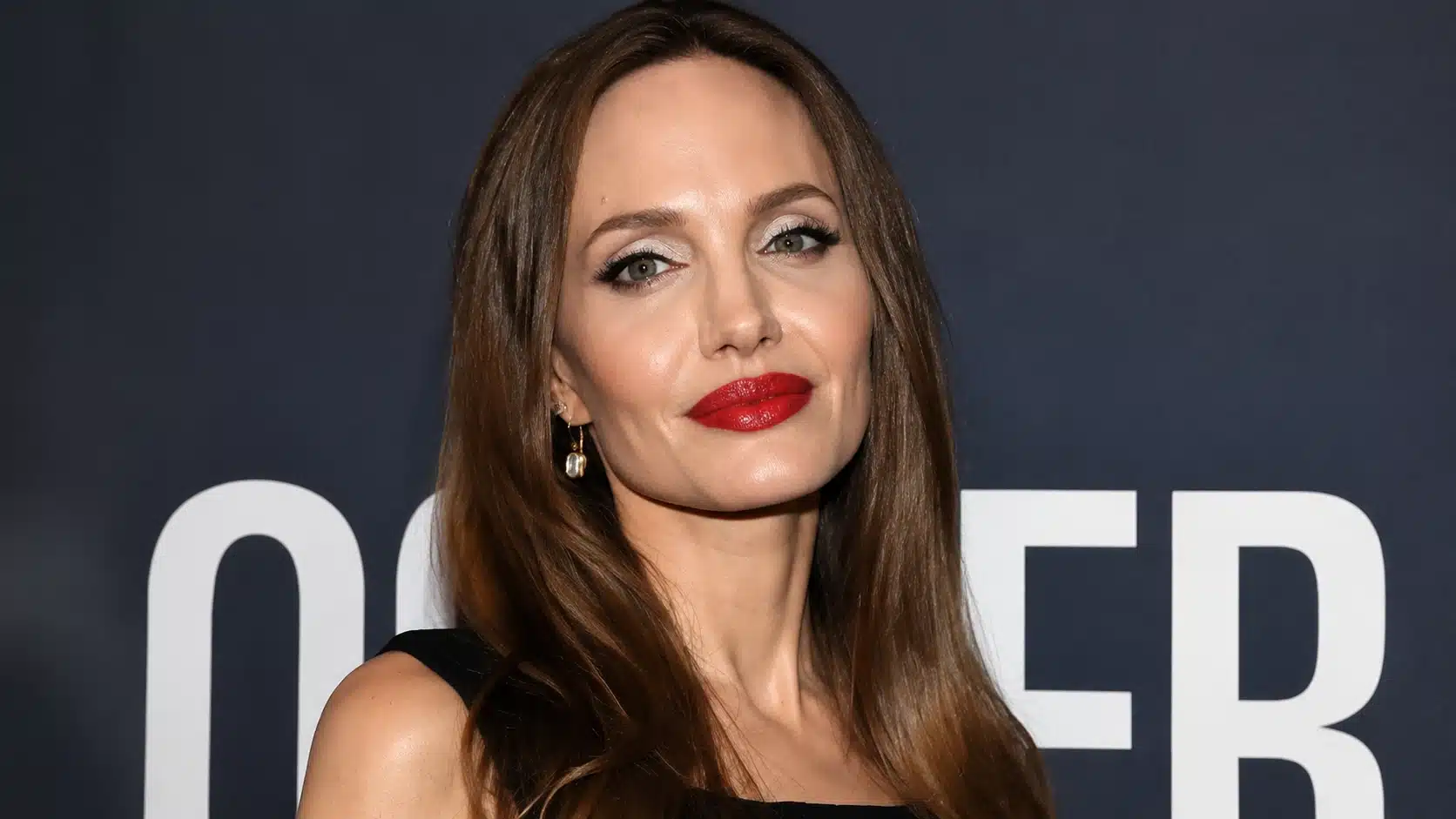 a woman with brown hair, wearing white eyeshadow and red lipstick, poses confidently against a dark backdrop