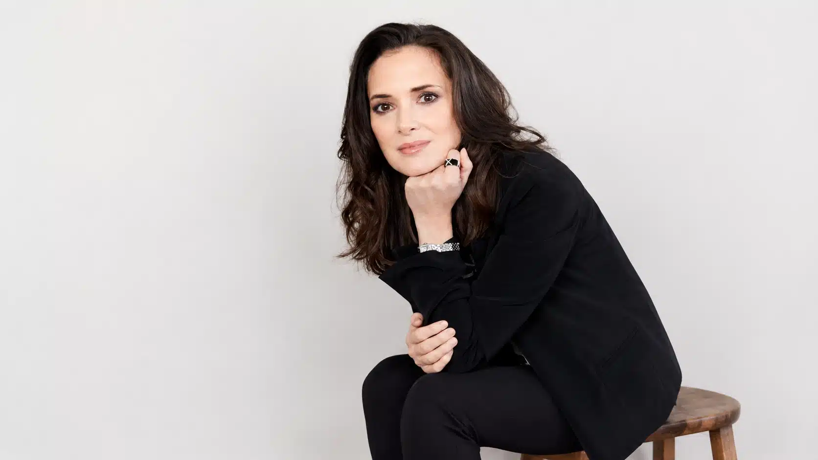 a woman with long dark hair, dressed in black, sits on a wooden stool with her hand on her chin