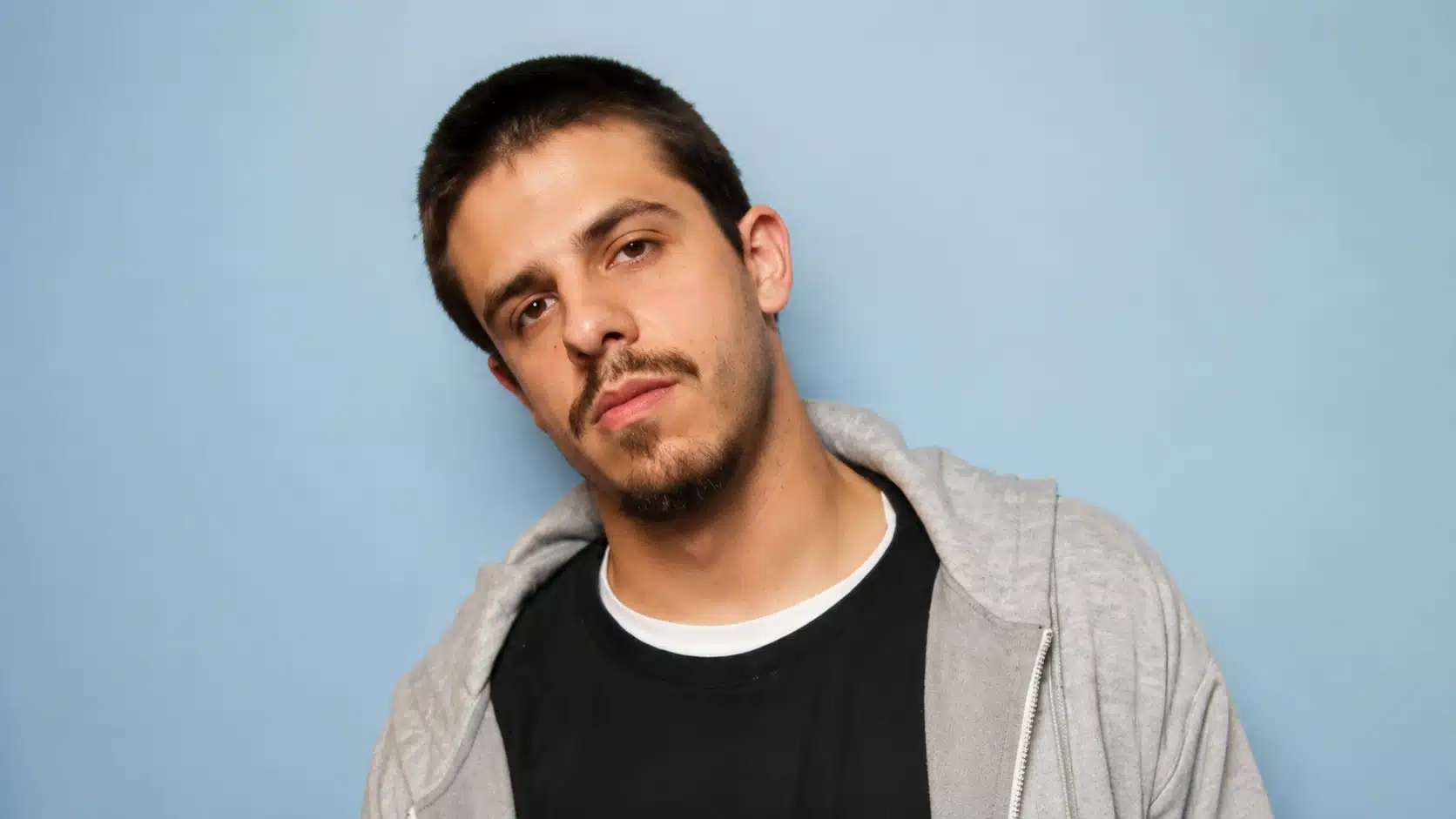 a young man in a gray hoodie and black t-shirt, standing against a light blue background with a serious expression