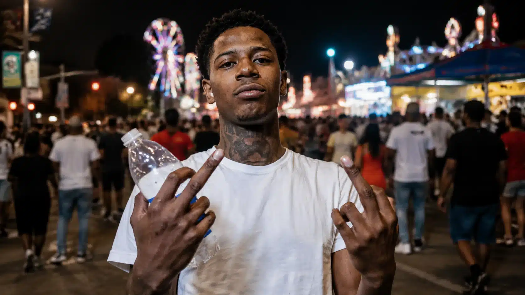 a young man in a white t-shirt making a peace sign, holding a water bottle, with a carnival backdrop at night