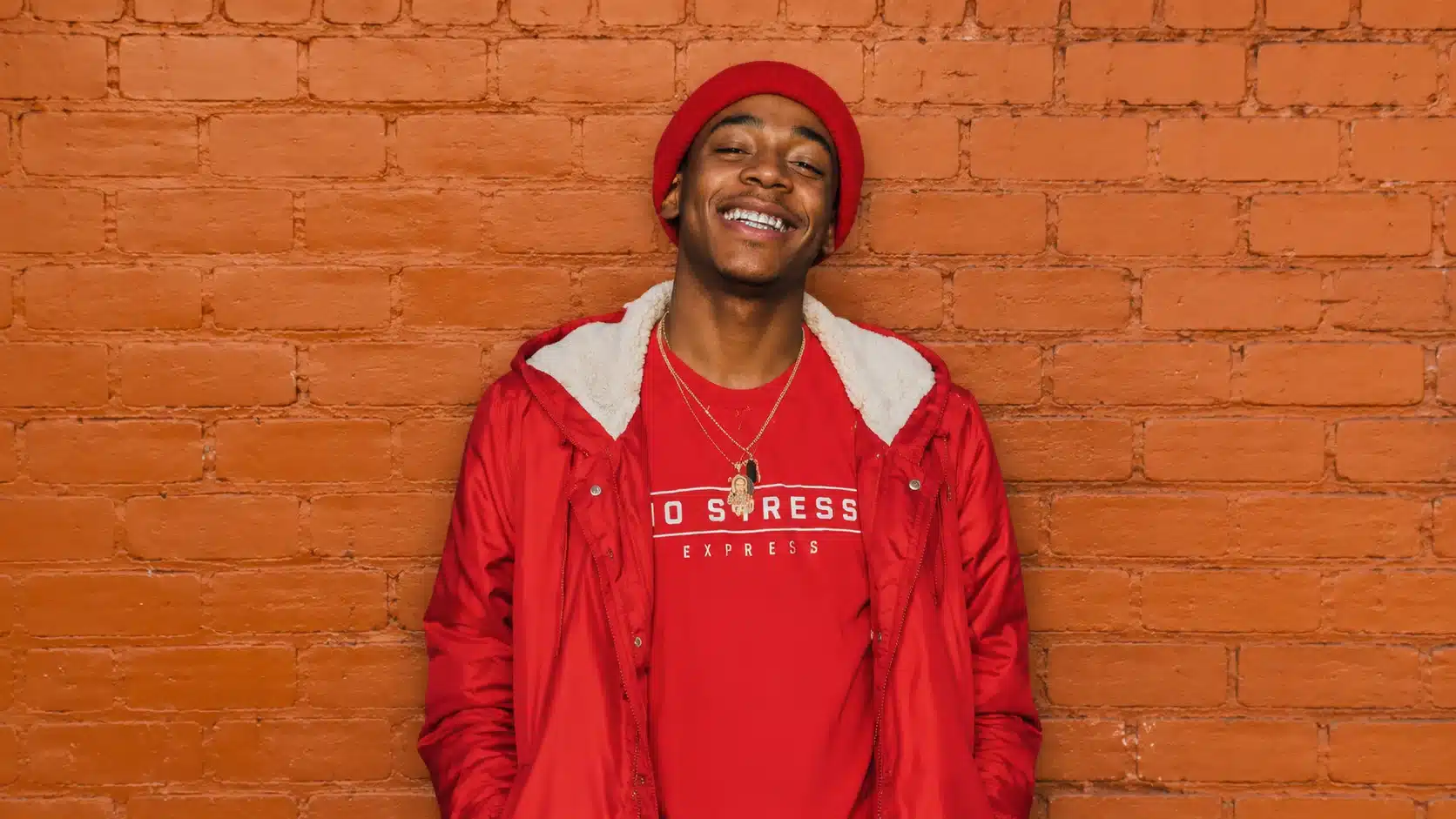 a young man wearing a red jacket and t-shirt, smiling in front of an orange brick wall with a confident pose