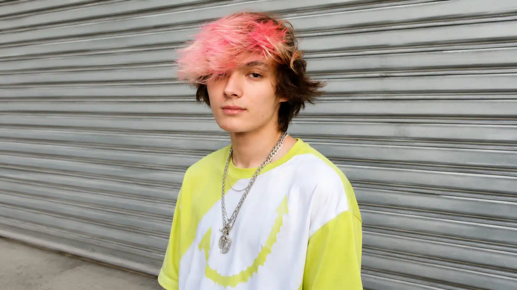 a young man with pink-highlighted hair, wearing a neon yellow and white shirt, standing in front of a metal gate