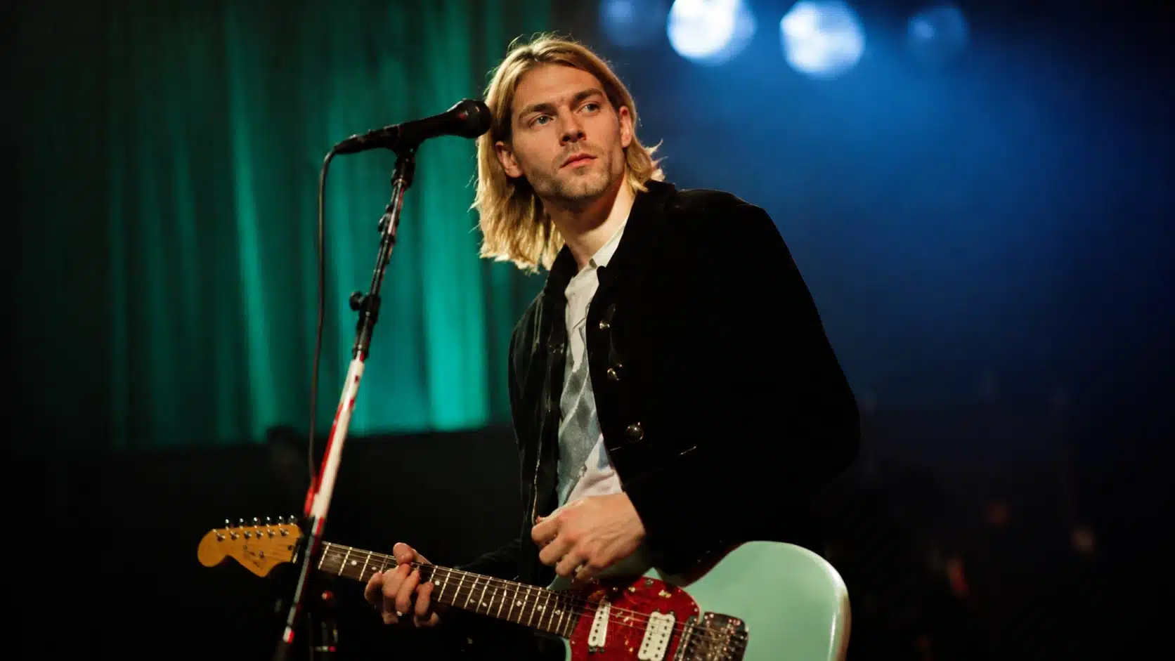 a young man with shoulder-length blonde hair playing a guitar on stage, looking towards the audience