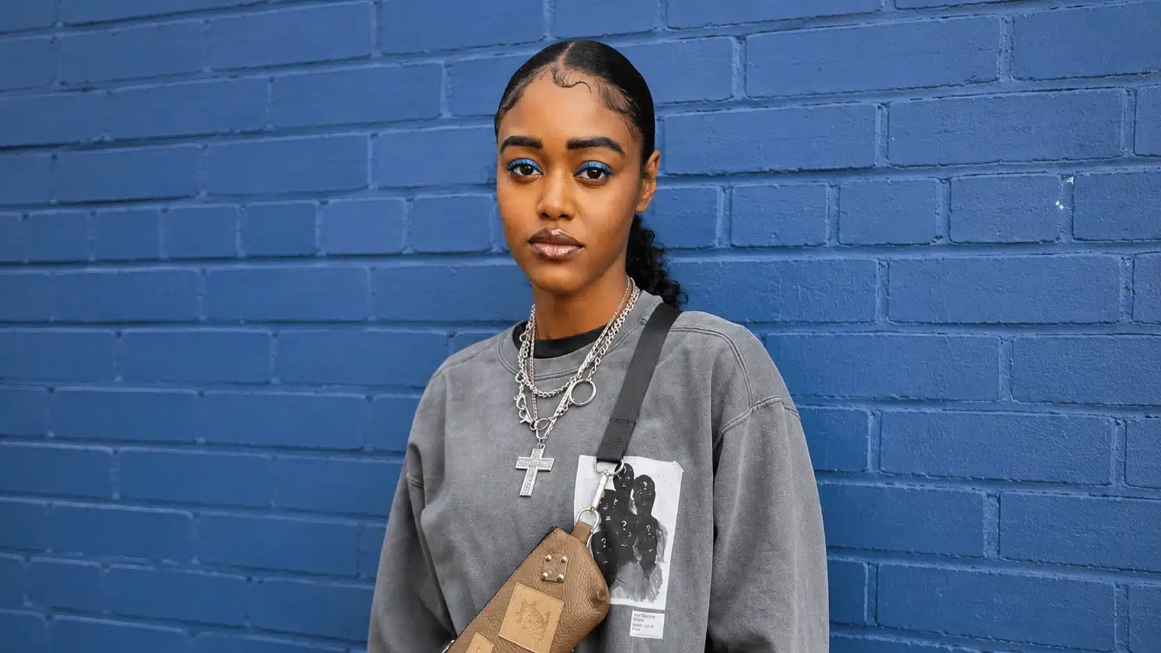 a young woman with blue eyeshadow wearing a gray sweatshirt and layered necklaces, standing in front of blue brick wall