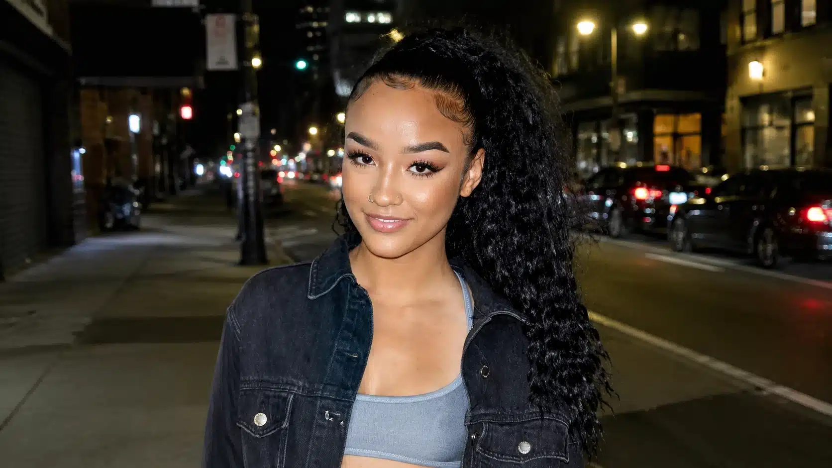 a young woman with curly hair smiling in a black denim jacket, standing on a city street at night