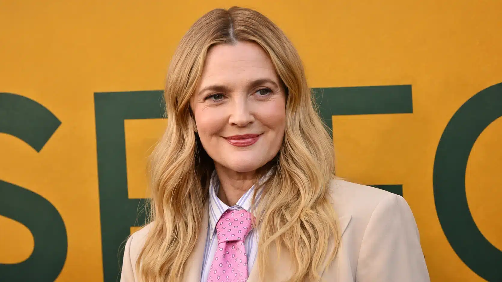 blonde woman in beige blazer and pink tie smiles softly, standing against mustard wall with green lettering