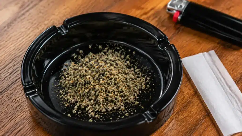 close-up of finely ground mimosa cannabis in a black ashtray on a wooden table, with rolling paper and lighter nearby