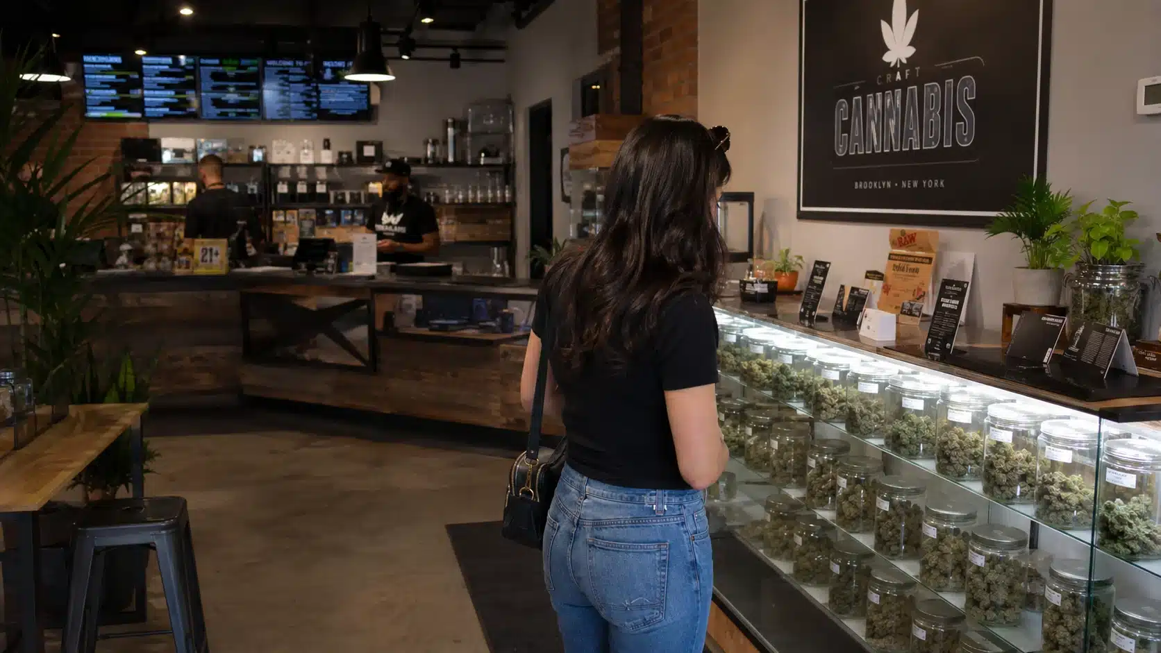 customer browsing cannabis jars in a modern dispensary, with glass display cases, staff, plants, and interior lighting