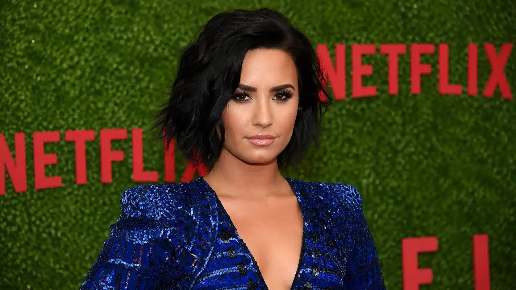 woman in a blue shimmery dress posing before a green backdrop with large red letters behind her