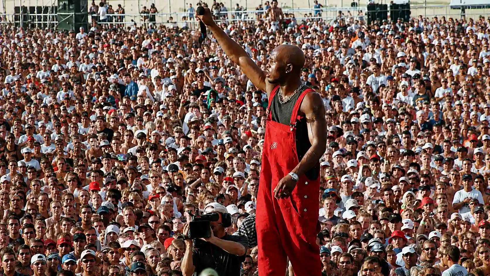 dmx in red overalls raises a microphone while performing in front of a large outdoor crowd.