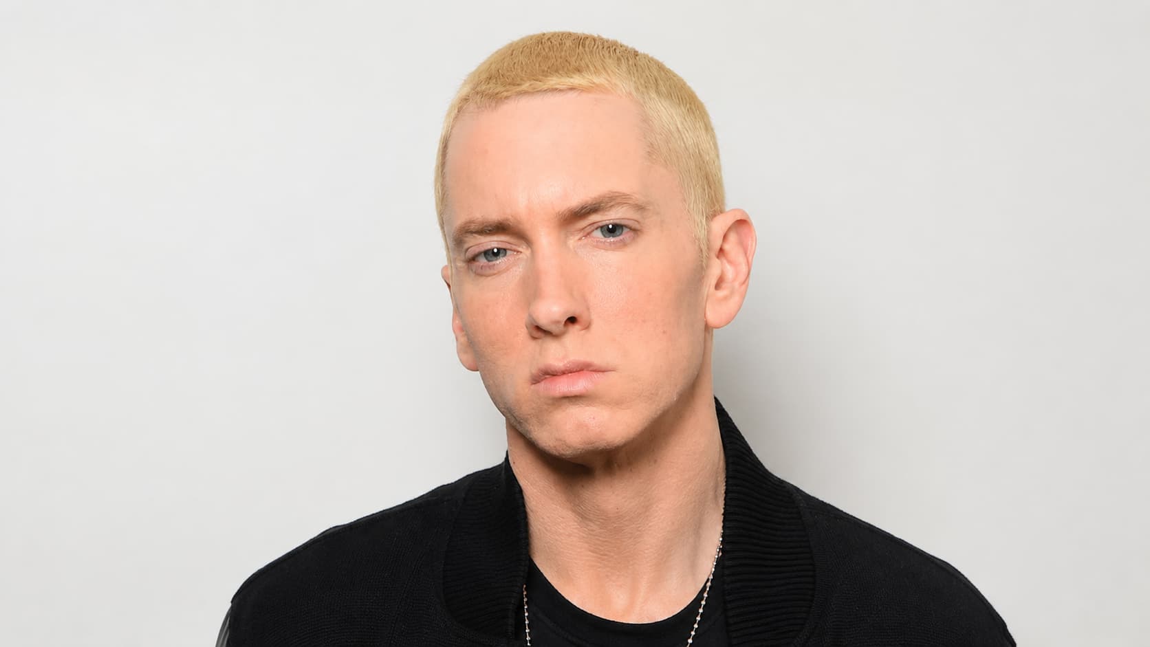 eminem with very short blond hair wearing a black jacket looking at camera with serious expression
