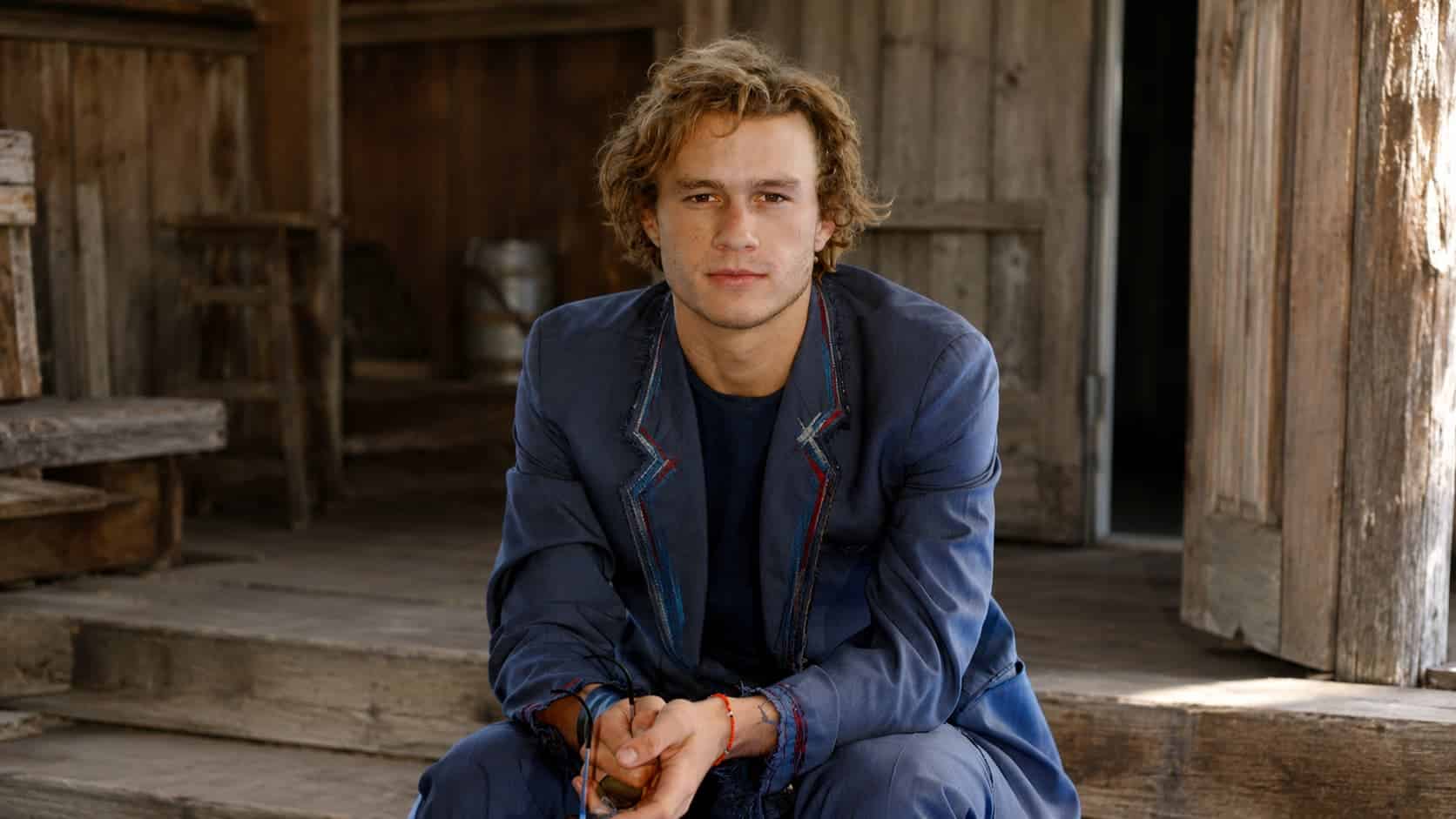 heath ledger in blue suit seated in a rustic wooden barn setting with calm face