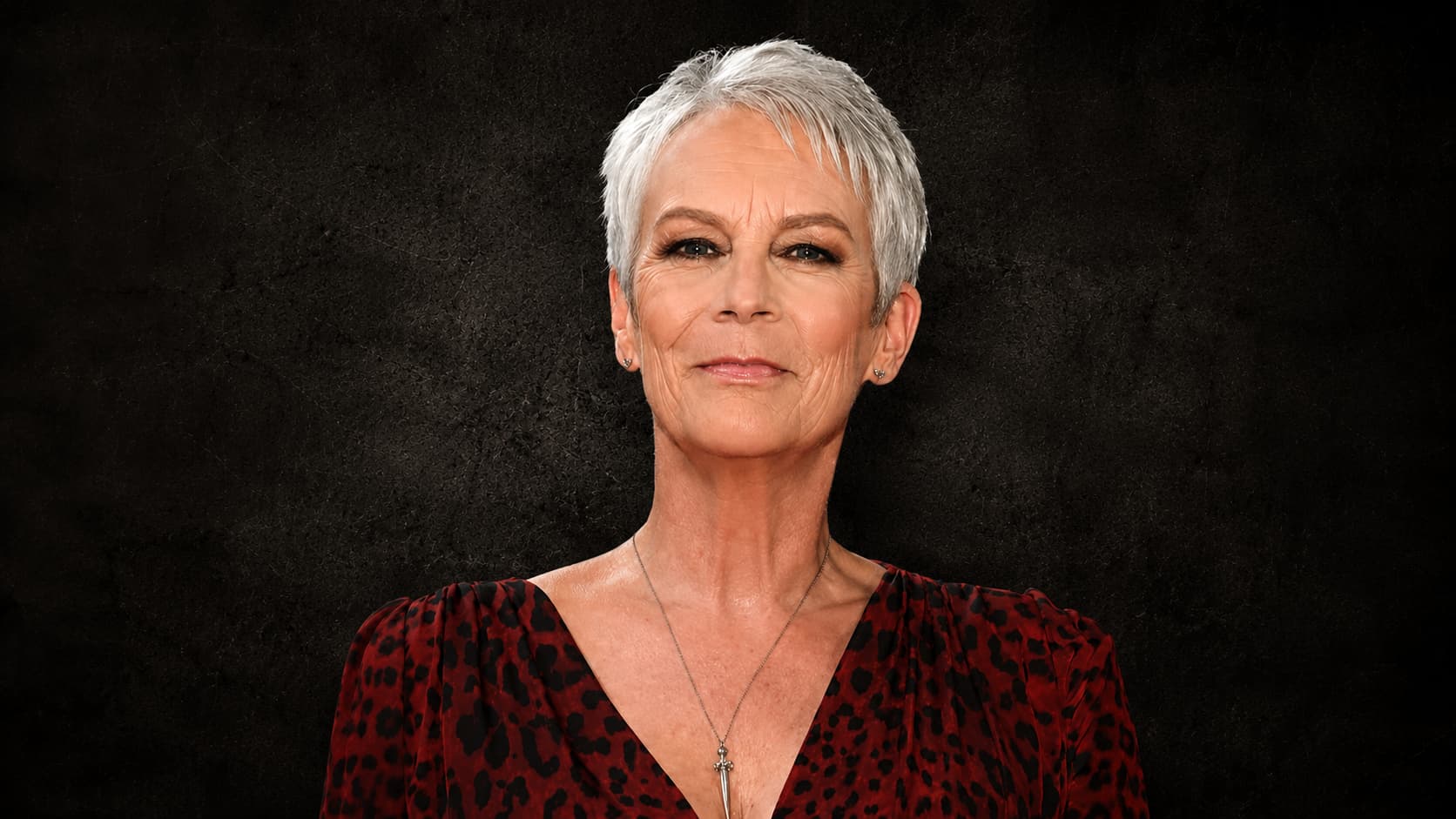Jamie Lee Curtis with short white hair, smiling at the camera against black rustic background