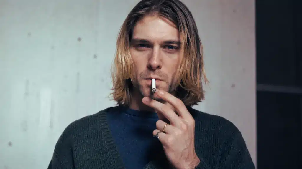 kurt cobain with shoulder-length blonde hair, wearing a green sweater, holding a cigarette and looking at the camera