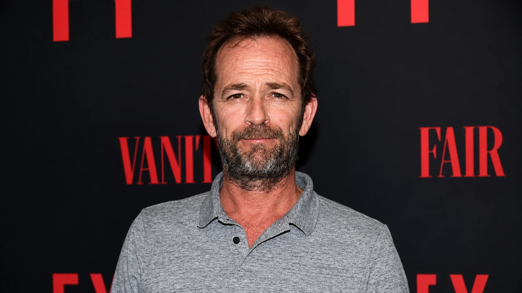 realistic portrait of a bearded man in a gray polo standing before a black backdrop with bold red event letters