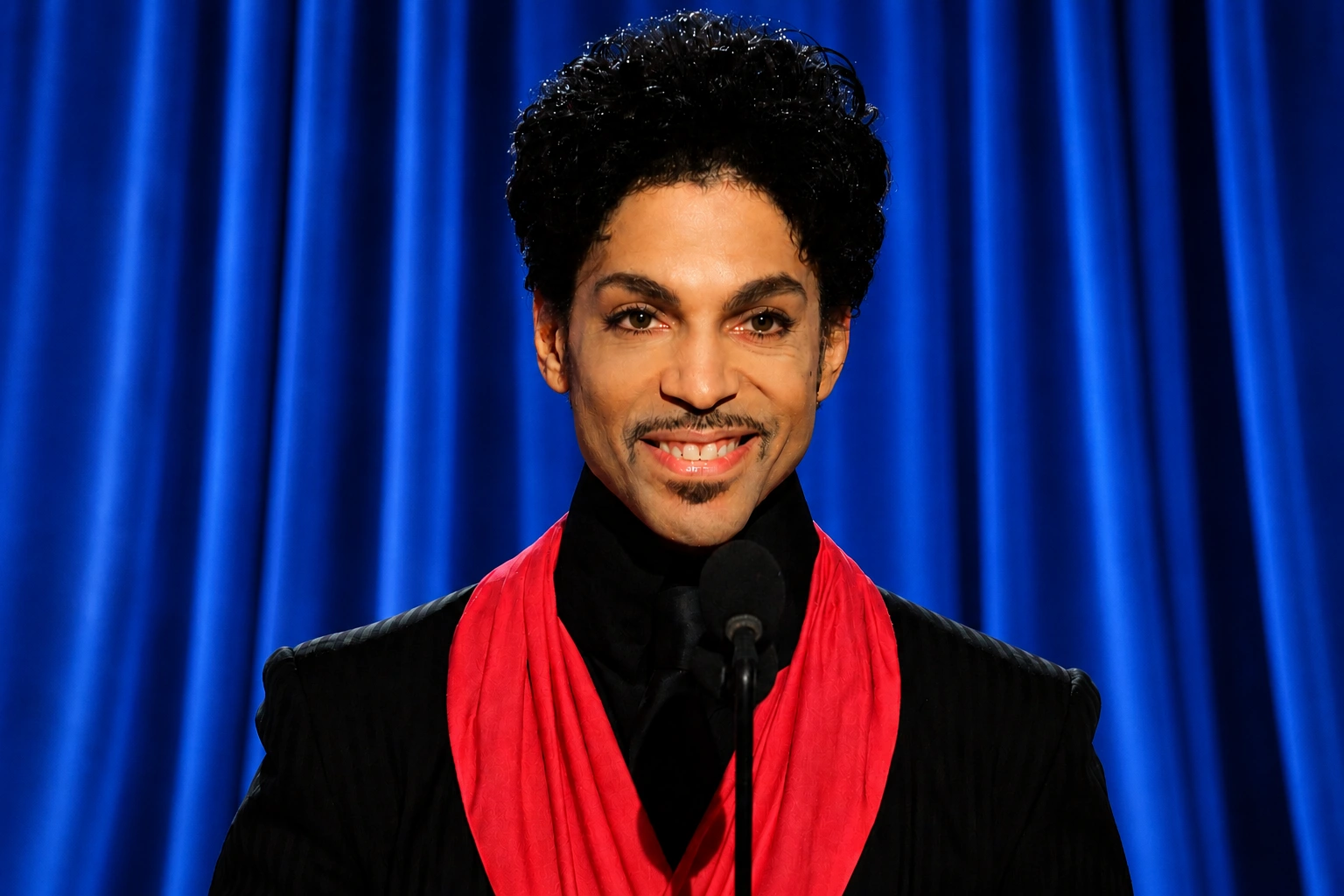 man wearing black outfit and red scarf speaking at microphone in front of deep blue curtain backdrop