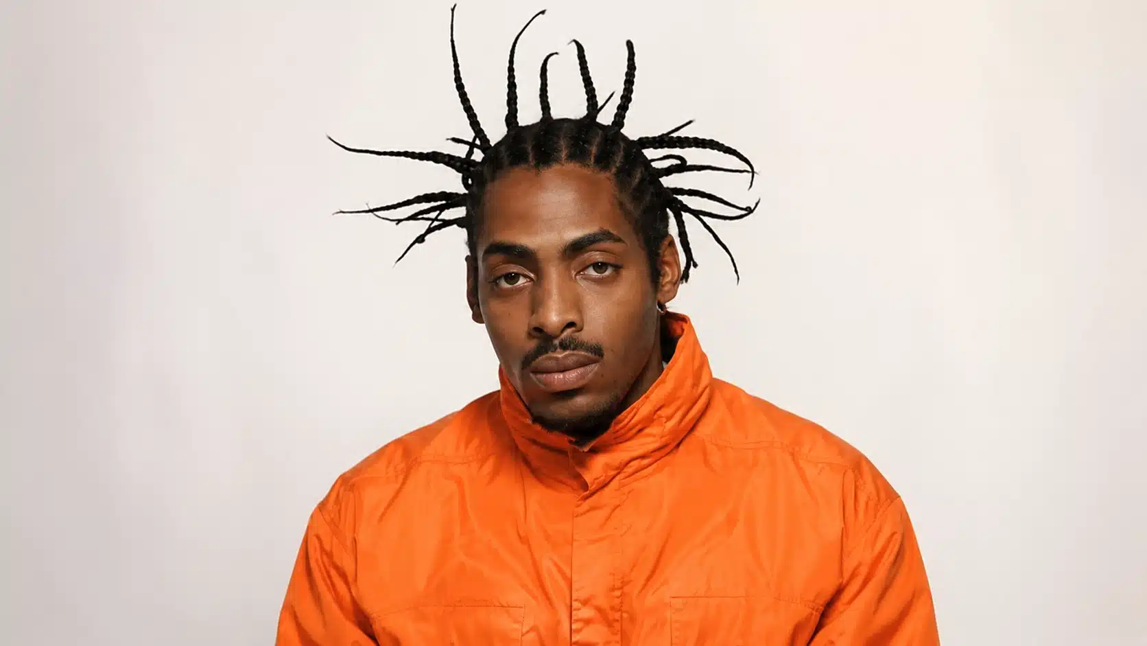 man with braided hair wearing an orange jacket, standing against a plain light background with a serious expression