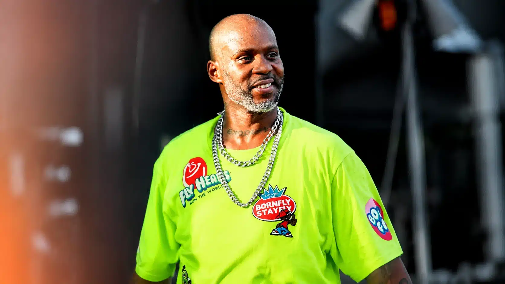 man with shaved head and silver beard wearing a neon green graphic t-shirt and chains, smiling on stage