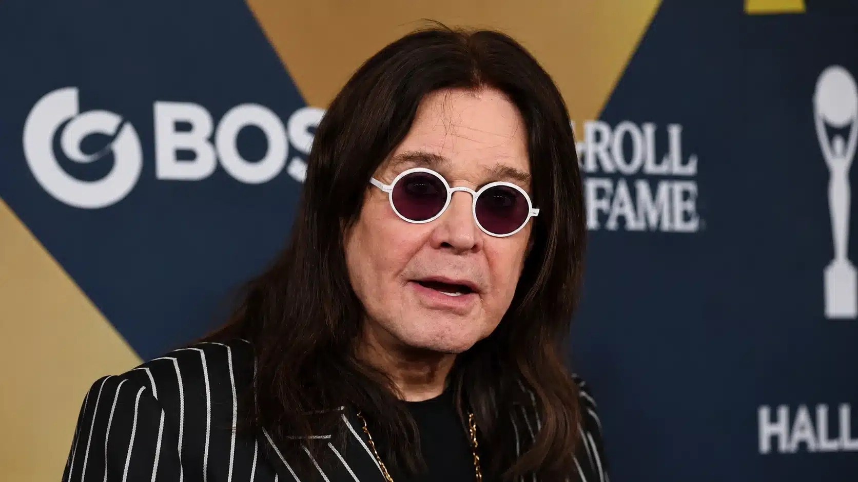 middle-aged man with long dark hair, wearing black pinstripe jacket and white sunglasses, posing at blue and gold wall