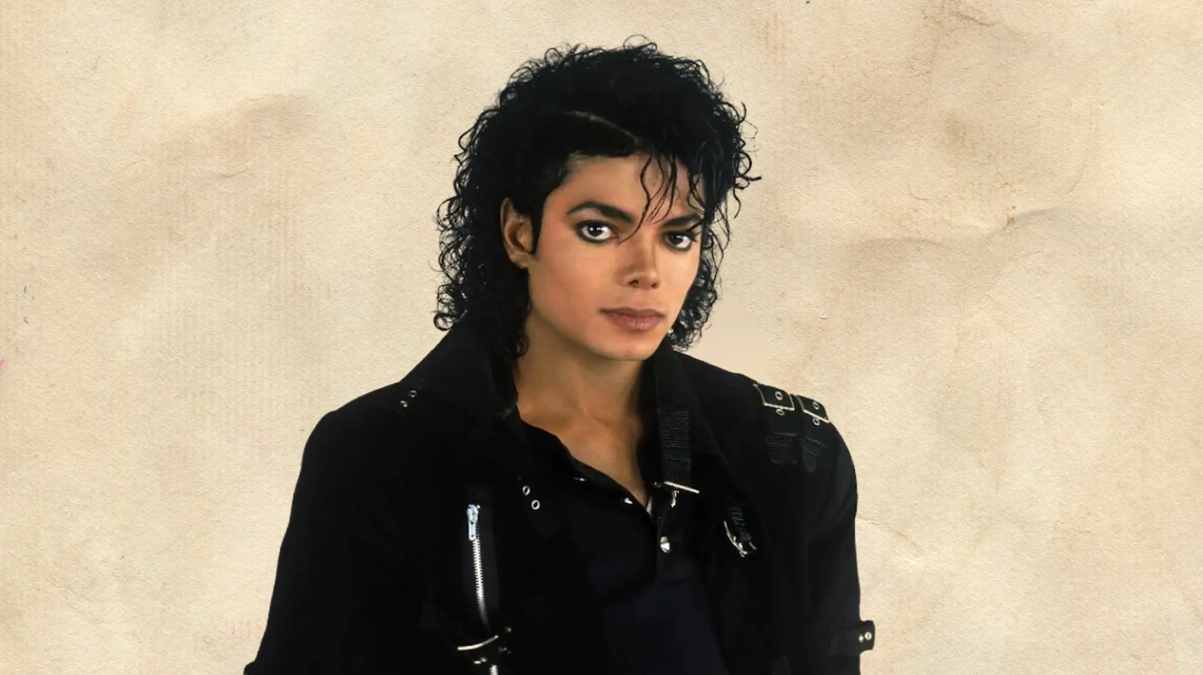 person with curly dark hair wearing black outfit, looking at camera with serious expression against beige background