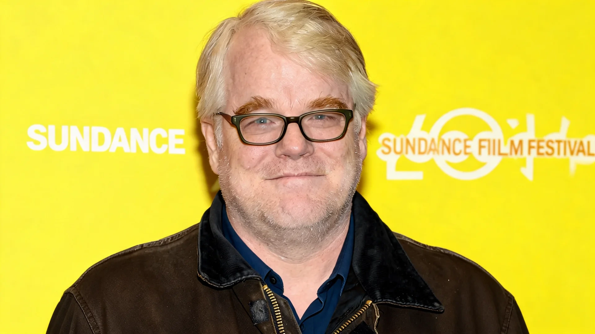 portrait of a man with glasses and white hair wearing a dark jacket against a bright yellow festival backdrop
