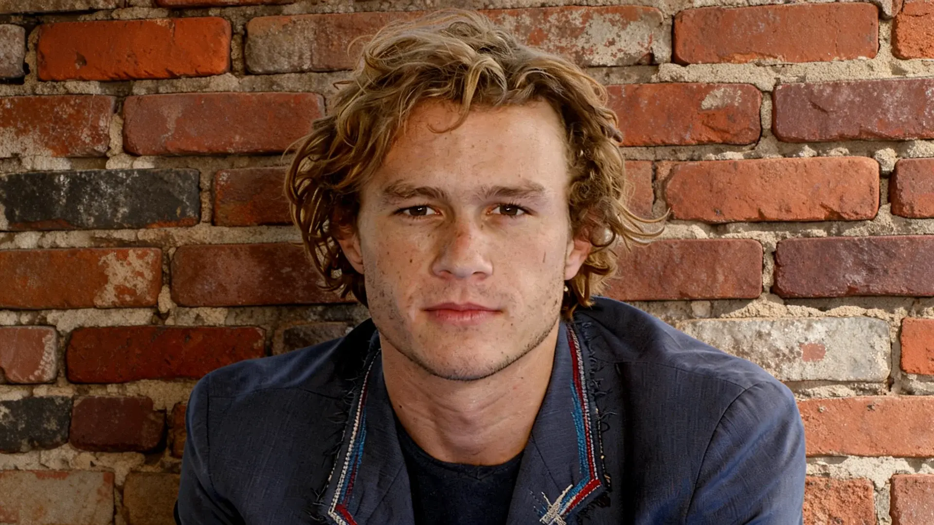 portrait of a young man with curly hair wearing a dark jacket while sitting in front of a red brick wall